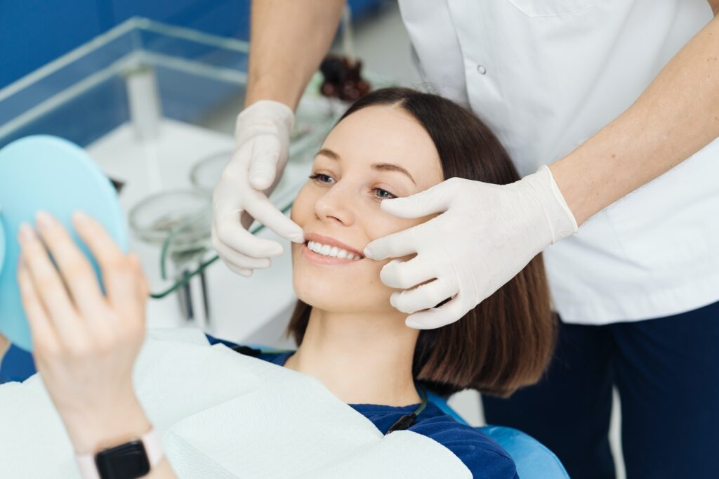 Female dental patient holding a mirror while the dentist examines her smile with gloved hands.