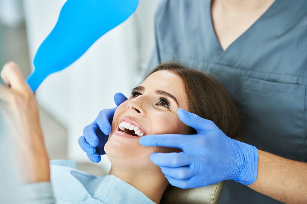 Young woman smiling while looking in a mirror during a dental exam with gloved hands checking her teeth.